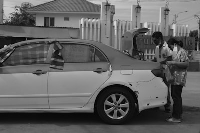 A black and white photo shows a sedan with visible damage and tape on the rear door. Two individuals are standing at the open trunk, engaged in arranging or examining its contents. The background features a building with a tiled roof and exposed piping.