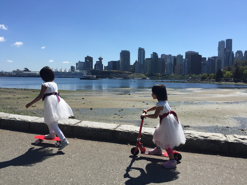 two little girls in dresses are riding scooters by the water