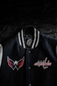 A black varsity jacket with white and gray striped collar is displayed against a dark background. The jacket features embroidered logos of a sports team, with a winged logo on the left side and the text 'Washington Capitals' on the right side, accompanied by three stars.