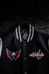 A black varsity jacket with white and gray striped collar is displayed against a dark background. The jacket features embroidered logos of a sports team, with a winged logo on the left side and the text 'Washington Capitals' on the right side, accompanied by three stars.
