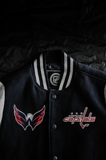 A black varsity jacket with white and gray striped collar is displayed against a dark background. The jacket features embroidered logos of a sports team, with a winged logo on the left side and the text 'Washington Capitals' on the right side, accompanied by three stars.