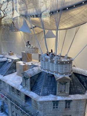 A detailed miniature scene featuring two wicker balloon baskets suspended over a snow-covered model of a building. The model depicts a European-style architecture with intricate details such as windows and shutters, enhanced with faux snow. Visible in the reflection, there are city buildings and trees in the glass window backdrop.