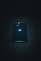 A sleek, dark-colored smartphone with a prominent logo positioned near the center. It features dual camera lenses on the top left corner of its backside. The phone is resting on a dark surface, and the overall lighting creates a moody, dramatic effect.