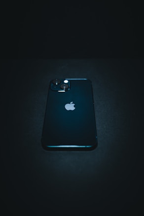 A sleek, dark-colored smartphone with a prominent logo positioned near the center. It features dual camera lenses on the top left corner of its backside. The phone is resting on a dark surface, and the overall lighting creates a moody, dramatic effect.