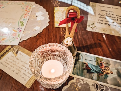 A glass candle holder with a burning candle is placed on a wooden table. Surrounding the candle are letters and decorative paper items. A small brass bell with a red ribbon and a decorative white flower is near the candle. There is also a religious illustration of a figure carrying another person on his back in a body of water.