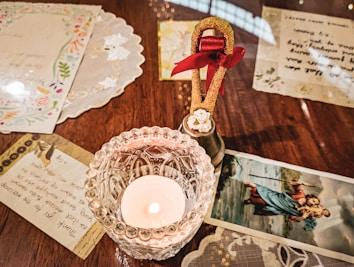 A glass candle holder with a burning candle is placed on a wooden table. Surrounding the candle are letters and decorative paper items. A small brass bell with a red ribbon and a decorative white flower is near the candle. There is also a religious illustration of a figure carrying another person on his back in a body of water.