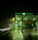 A dimly lit gas station during night time with a green and white color scheme. The station features an illuminated BP logo and a fuel price display, with multiple street lights providing additional lighting. The wet pavement suggests recent rainfall, reflecting the lights. There are no visible vehicles or people in the image.