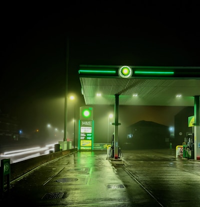 A dimly lit gas station during night time with a green and white color scheme. The station features an illuminated BP logo and a fuel price display, with multiple street lights providing additional lighting. The wet pavement suggests recent rainfall, reflecting the lights. There are no visible vehicles or people in the image.