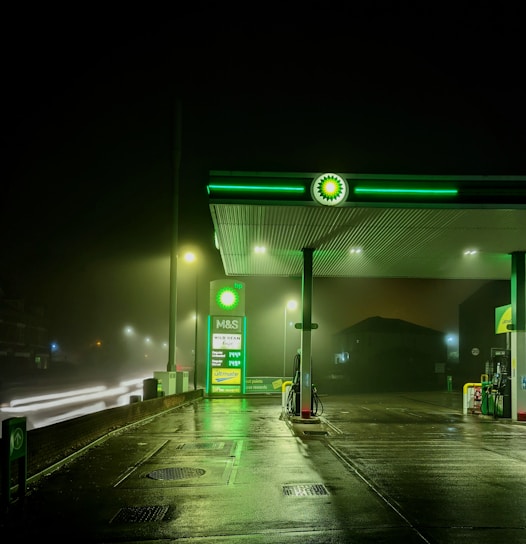 A dimly lit gas station during night time with a green and white color scheme. The station features an illuminated BP logo and a fuel price display, with multiple street lights providing additional lighting. The wet pavement suggests recent rainfall, reflecting the lights. There are no visible vehicles or people in the image.