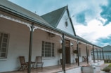 A charming house with a gabled roof and ornate white trim, featuring a long veranda with wooden chairs. The sky is cloudy, giving a dramatic backdrop to the classic architecture. Potted plants hang near the entrance, adding a touch of greenery.
