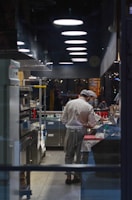 A chef in a well-equipped, modern kitchen is preparing food on a countertop. Overhead lighting fixtures line the ceiling, and various kitchen equipment, including ovens and shelves with condiments, are neatly organized. The dimly lit kitchen creates a focused atmosphere with reflections visible in the glass window separating the kitchen from the dining area.
