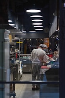 A chef in a well-equipped, modern kitchen is preparing food on a countertop. Overhead lighting fixtures line the ceiling, and various kitchen equipment, including ovens and shelves with condiments, are neatly organized. The dimly lit kitchen creates a focused atmosphere with reflections visible in the glass window separating the kitchen from the dining area.
