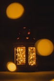 A lantern with intricate star-shaped cutouts glows softly, filled with delicate string lights that cast a warm, golden hue. Out-of-focus circular bokeh lights of similar color float around, enhancing the dreamy atmosphere.
