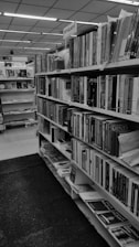 A library interior featuring a row of bookshelves filled with books. The sign above one section reads 'Mythology'. The shelves contain various books of different sizes and thicknesses, arranged in an organized manner. The space is well-lit, and the library seems spacious and orderly.