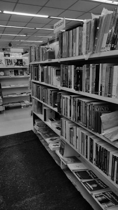 A library interior featuring a row of bookshelves filled with books. The sign above one section reads 'Mythology'. The shelves contain various books of different sizes and thicknesses, arranged in an organized manner. The space is well-lit, and the library seems spacious and orderly.