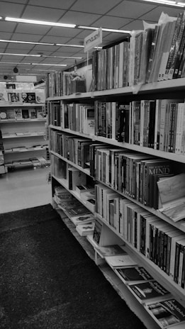 A library interior featuring a row of bookshelves filled with books. The sign above one section reads 'Mythology'. The shelves contain various books of different sizes and thicknesses, arranged in an organized manner. The space is well-lit, and the library seems spacious and orderly.