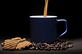 A dark blue mug is being filled with coffee, surrounded by roasted coffee beans and several cookies arranged neatly. The scene is set against a black background, creating a focus on the mug and its contents.