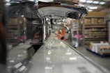 A close-up view of a row of shiny chrome faucets lined up on a display, with a warehouse setting in the background. The focus is on the faucets, emphasizing their metallic finish and curved design, while shelves filled with boxes and equipment can be seen blurred in the distance.