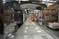 A close-up view of a row of shiny chrome faucets lined up on a display, with a warehouse setting in the background. The focus is on the faucets, emphasizing their metallic finish and curved design, while shelves filled with boxes and equipment can be seen blurred in the distance.