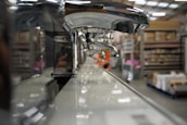A close-up view of a row of shiny chrome faucets lined up on a display, with a warehouse setting in the background. The focus is on the faucets, emphasizing their metallic finish and curved design, while shelves filled with boxes and equipment can be seen blurred in the distance.