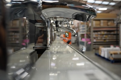 A close-up view of a row of shiny chrome faucets lined up on a display, with a warehouse setting in the background. The focus is on the faucets, emphasizing their metallic finish and curved design, while shelves filled with boxes and equipment can be seen blurred in the distance.