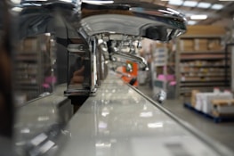 A close-up view of a row of shiny chrome faucets lined up on a display, with a warehouse setting in the background. The focus is on the faucets, emphasizing their metallic finish and curved design, while shelves filled with boxes and equipment can be seen blurred in the distance.