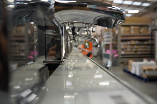 A close-up view of a row of shiny chrome faucets lined up on a display, with a warehouse setting in the background. The focus is on the faucets, emphasizing their metallic finish and curved design, while shelves filled with boxes and equipment can be seen blurred in the distance.