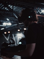 A person is wearing a virtual reality headset and holding a device with a circular light feature. The setting appears to be a dimly lit technological or industrial environment, with exposed metal beams and soft lighting.