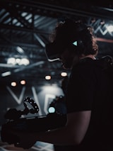 A person is wearing a virtual reality headset and holding a device with a circular light feature. The setting appears to be a dimly lit technological or industrial environment, with exposed metal beams and soft lighting.