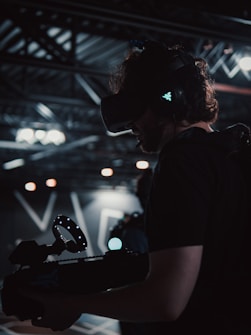 A person is wearing a virtual reality headset and holding a device with a circular light feature. The setting appears to be a dimly lit technological or industrial environment, with exposed metal beams and soft lighting.