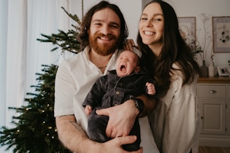 A couple is smiling and posing indoors while holding a yawning baby. They are standing beside a decorated Christmas tree. The man has a beard and long hair, while the woman has long straight hair. The background includes a sideboard with decorative items and framed pictures on the wall.