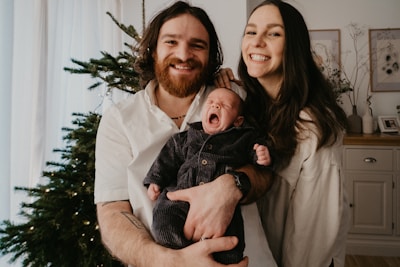 A couple is smiling and posing indoors while holding a yawning baby. They are standing beside a decorated Christmas tree. The man has a beard and long hair, while the woman has long straight hair. The background includes a sideboard with decorative items and framed pictures on the wall.