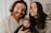 A cheerful family portrait featuring a bearded man with long hair smiling widely, a woman with straight long hair making a playful expression, and a baby with a curious look. They are all dressed in casual attire with light-colored backgrounds suggesting an indoor setting.