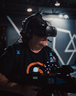 A person immersed in virtual reality, wearing a VR headset and holding a futuristic-looking toy gun with LED lights. The background features geometric patterns with a dimly lit atmosphere, evoking a high-tech gaming environment.
