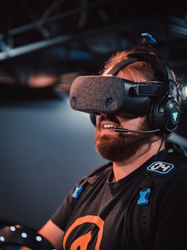A person is wearing a virtual reality headset and headphones. They have a beard and are dressed in casual clothing with a logo on the shirt. The background is dimly lit, suggesting an indoor environment, possibly a gaming or tech setting.
