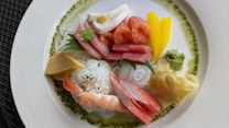 A beautifully arranged plate of assorted sashimi, featuring slices of fresh salmon and tuna, alongside crab sticks, shrimp, and a variety of garnishes including wasabi, ginger, and a mound of white rice topped with sesame seeds. The dish is accented with vibrant yellow mango slices and a green leaf, all artistically presented on a white plate with a green sauce ring around the edge.