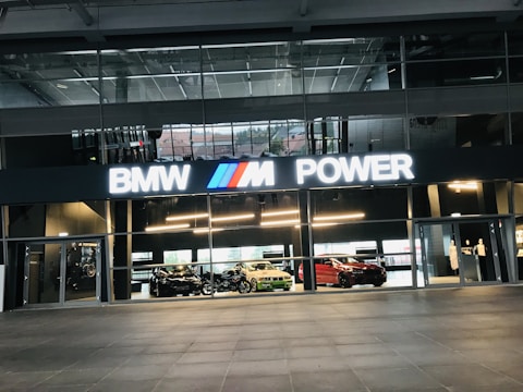 A modern building with large glass windows and a bold BMW M Power sign illuminated above a showroom entrance. Inside, several BMW cars are on display, including a black, white, and red car. The interior has sleek lines and glass partitions, suggesting a luxurious and high-tech environment.