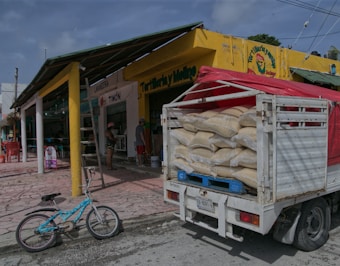 A small street scene with a truck loaded with large sacks parked in front of a yellow building with signage. A bicycle is resting against the curb, and a few people can be seen near the entrance of a storefront. The ground is paved with pinkish stone tiles, and the sky is overcast.