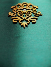 An ornate, symmetrical wooden decoration with intricate patterns sits on a textured teal background. The design features swirling lines and curves, giving it an elegant and artistic appearance.