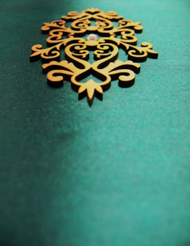 An ornate, symmetrical wooden decoration with intricate patterns sits on a textured teal background. The design features swirling lines and curves, giving it an elegant and artistic appearance.