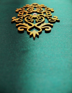 An ornate, symmetrical wooden decoration with intricate patterns sits on a textured teal background. The design features swirling lines and curves, giving it an elegant and artistic appearance.