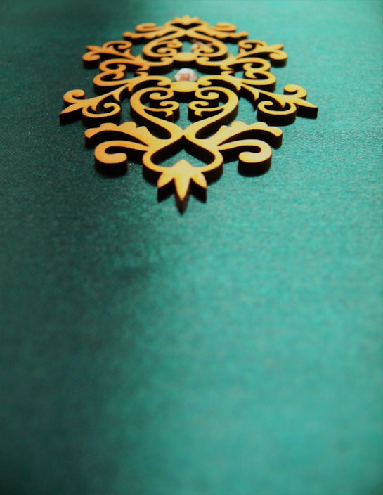An ornate, symmetrical wooden decoration with intricate patterns sits on a textured teal background. The design features swirling lines and curves, giving it an elegant and artistic appearance.