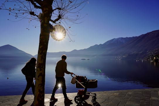 Silhouettes of two people walking along a lakeside path with a stroller, set against the backdrop of mountains and a calm lake. A nearly bare tree stands in the foreground, with the sun shining through its branches, creating a soft glow. The distant mountains are partially snow-capped, and the sky is clear, giving the scene a serene, peaceful atmosphere.
