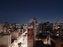 A nighttime cityscape featuring a densely packed array of tall buildings, the scene captures the calm and quiet of an urban environment under a starlit sky. In the foreground, a prominent mural depicts a human face with the word 'veracidade' visible, suggesting themes of truth or authenticity. The streets below are brightly lit with streetlights and light trails from passing vehicles, indicating motion and life even in the stillness of night.