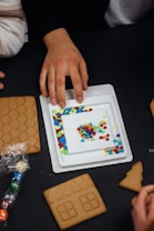 A hand reaches towards a tray filled with colorful candies, such as blue, red, yellow, and green. Surrounding the tray are gingerbread cookie pieces, including a roof tile design and a house shape with window outlines. Additional candies are seen in a plastic wrapper on a dark surface.