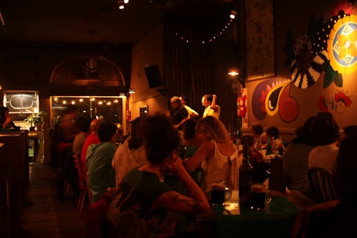 A dimly lit, cozy bar or restaurant with patrons seated at tables. There is warm, ambient lighting and colorful murals on the wall. A few people seem to be having a conversation, and drinks are placed on the tables.