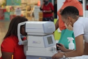A person is using an eye examination machine with the assistance of another individual. The setting appears to be indoors, possibly in a clinic or a mall. The equipment is white and has a small screen and various controls. Surrounding the scene are people and various items in the background.