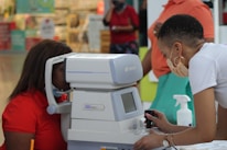 A person is using an eye examination machine with the assistance of another individual. The setting appears to be indoors, possibly in a clinic or a mall. The equipment is white and has a small screen and various controls. Surrounding the scene are people and various items in the background.