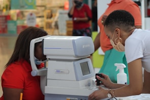 A person is using an eye examination machine with the assistance of another individual. The setting appears to be indoors, possibly in a clinic or a mall. The equipment is white and has a small screen and various controls. Surrounding the scene are people and various items in the background.