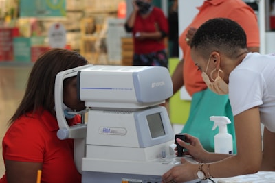 A person is using an eye examination machine with the assistance of another individual. The setting appears to be indoors, possibly in a clinic or a mall. The equipment is white and has a small screen and various controls. Surrounding the scene are people and various items in the background.